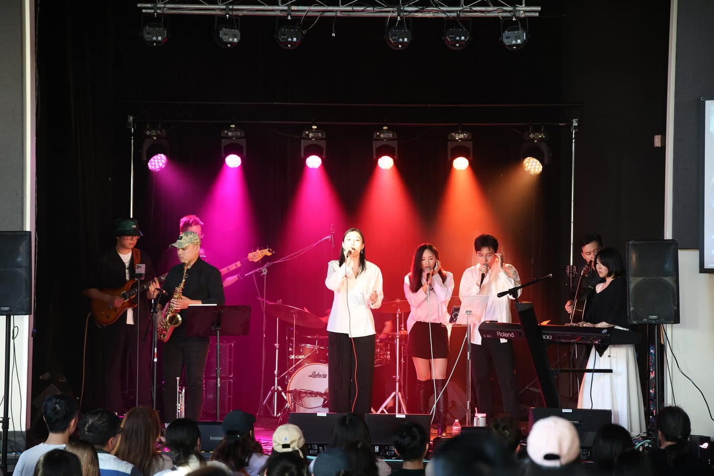 Full band performing on stage under a warm pink and amber light wash with the audience in view
