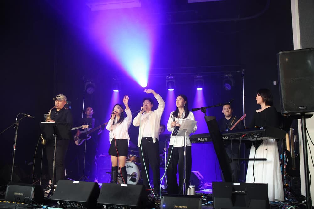 Full band ensemble raising their hands under a violet light wash